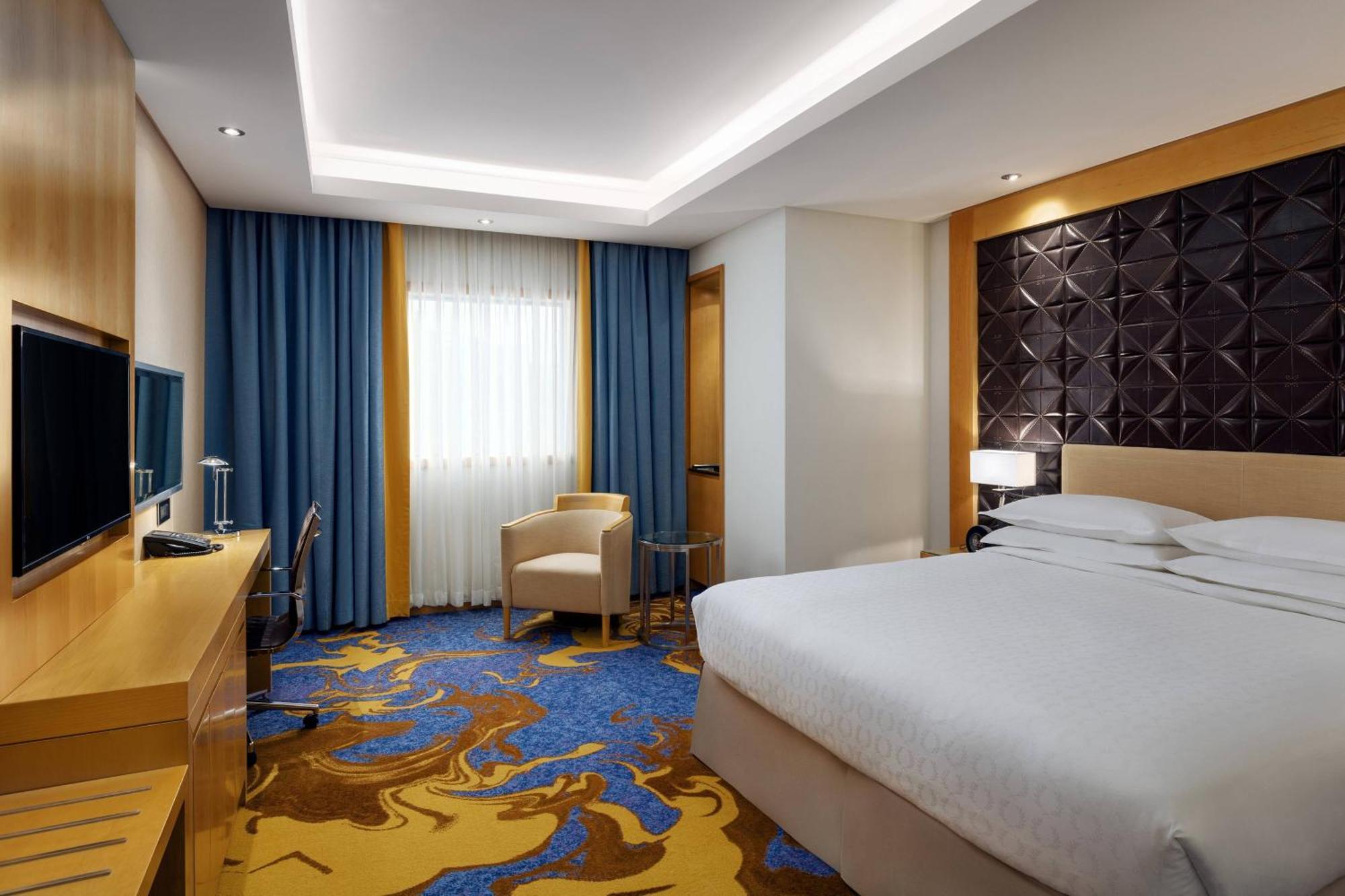 Sheraton Makkah Jabal Al Kaaba Hotel Mecca Exterior foto The photo shows a modern hotel room featuring a comfortable double bed with a light-colored headboard. There's a large window dressed with blue curtains, allowing natural light to enter. The walls are painted in neutral tones, and one wall has a text